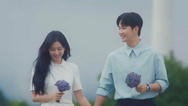 Dua tokoh utama drama Korea Queen of Tears berjalan
             berdampingan di bawah langit abu-abu yang tenang,
             masing-masing memegang buket bunga lavender kecil,
             ekspresi hangat dan penuh ketulusan